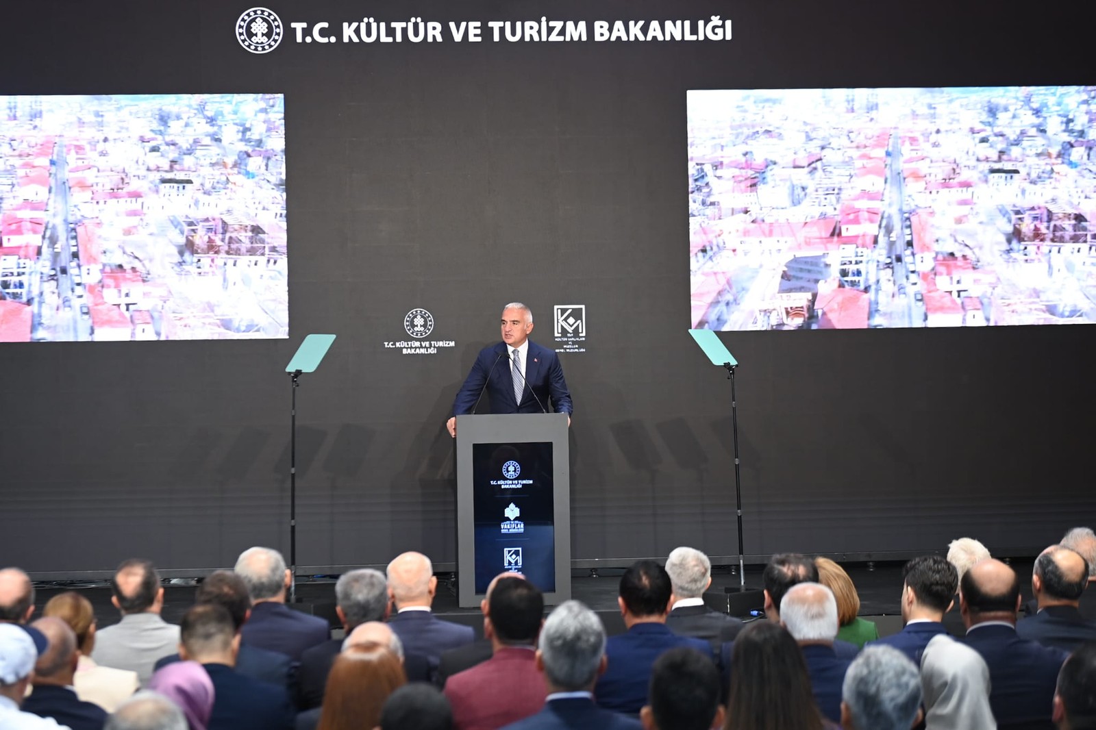 Bakanlık fonu önünde kürsüde konuşan yetkili; ekranlarda tarihi dokunun havadan görüntüsü