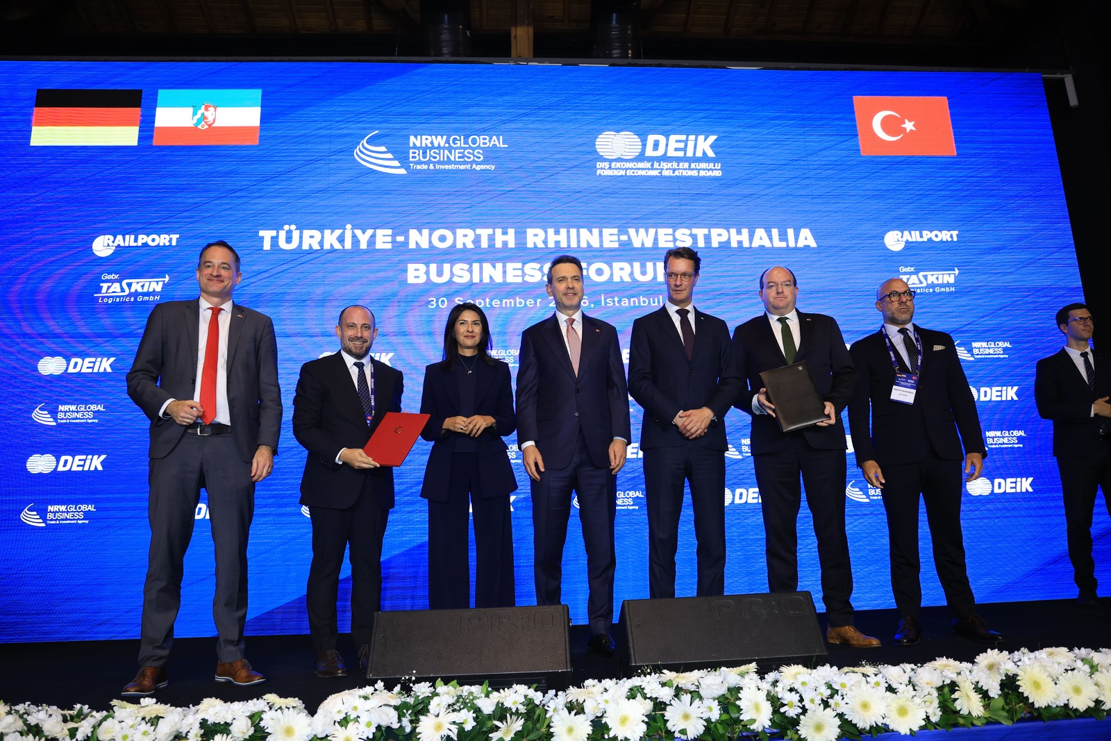 İş forumu sahnesinde bayraklar ve logolu ekran önünde, imza klasörleriyle toplu poz veren katılımcılar