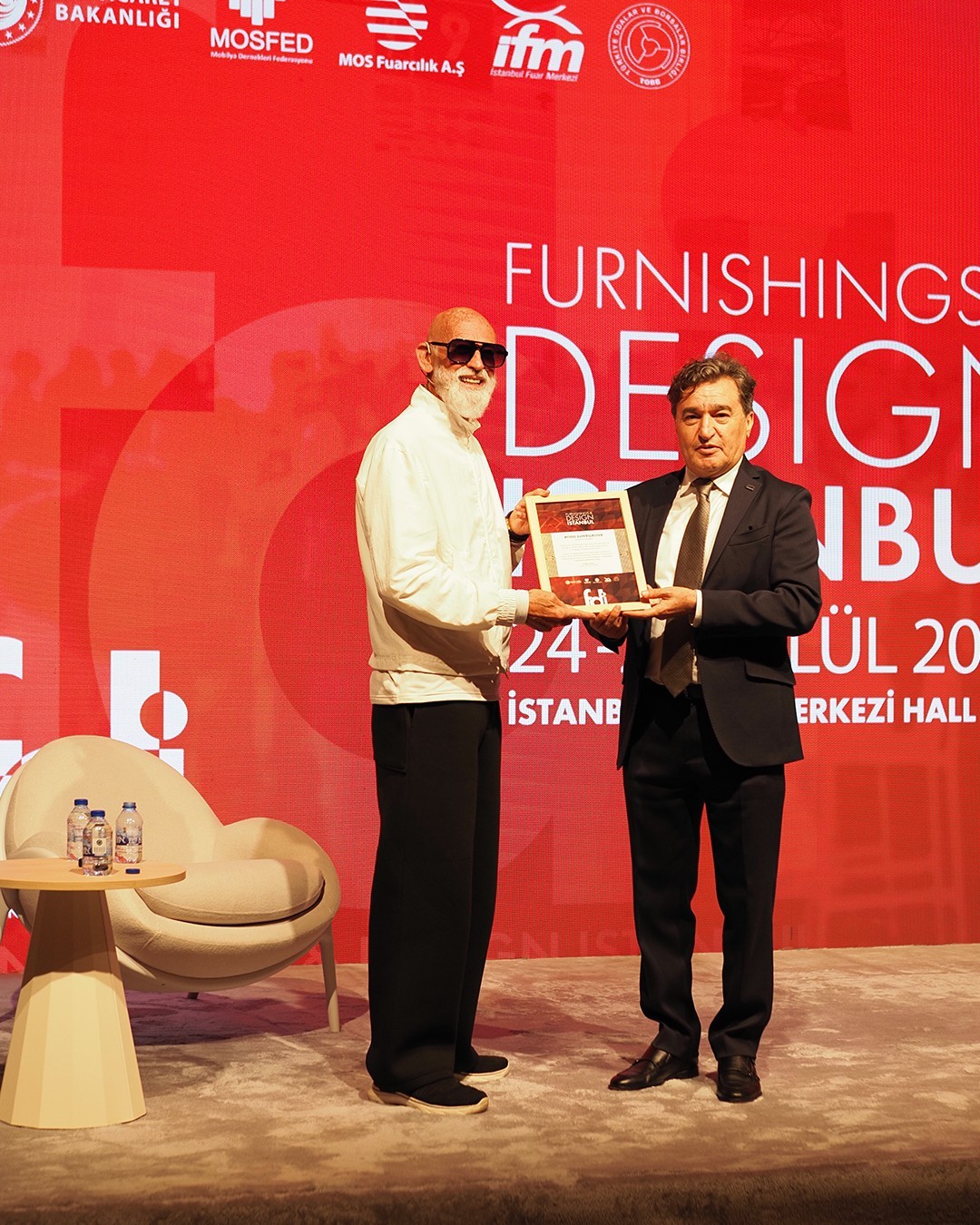 Furnishings Design İstanbul yazılı kırmızı sahne fonu önünde iki adam çerçeveli bir plaketi birlikte tutuyor