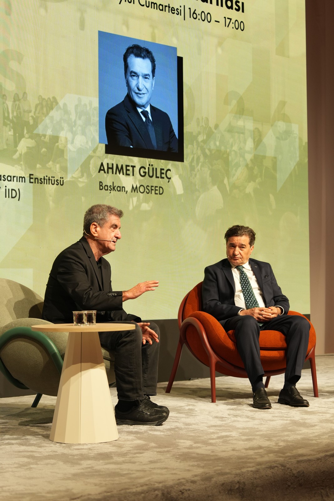 Fuar sahnesinde turuncu ve yeşil koltuklarda oturan iki erkek konuşmacı, arkada MOSFED Başkanı Ahmet Güleç yazılı ekran