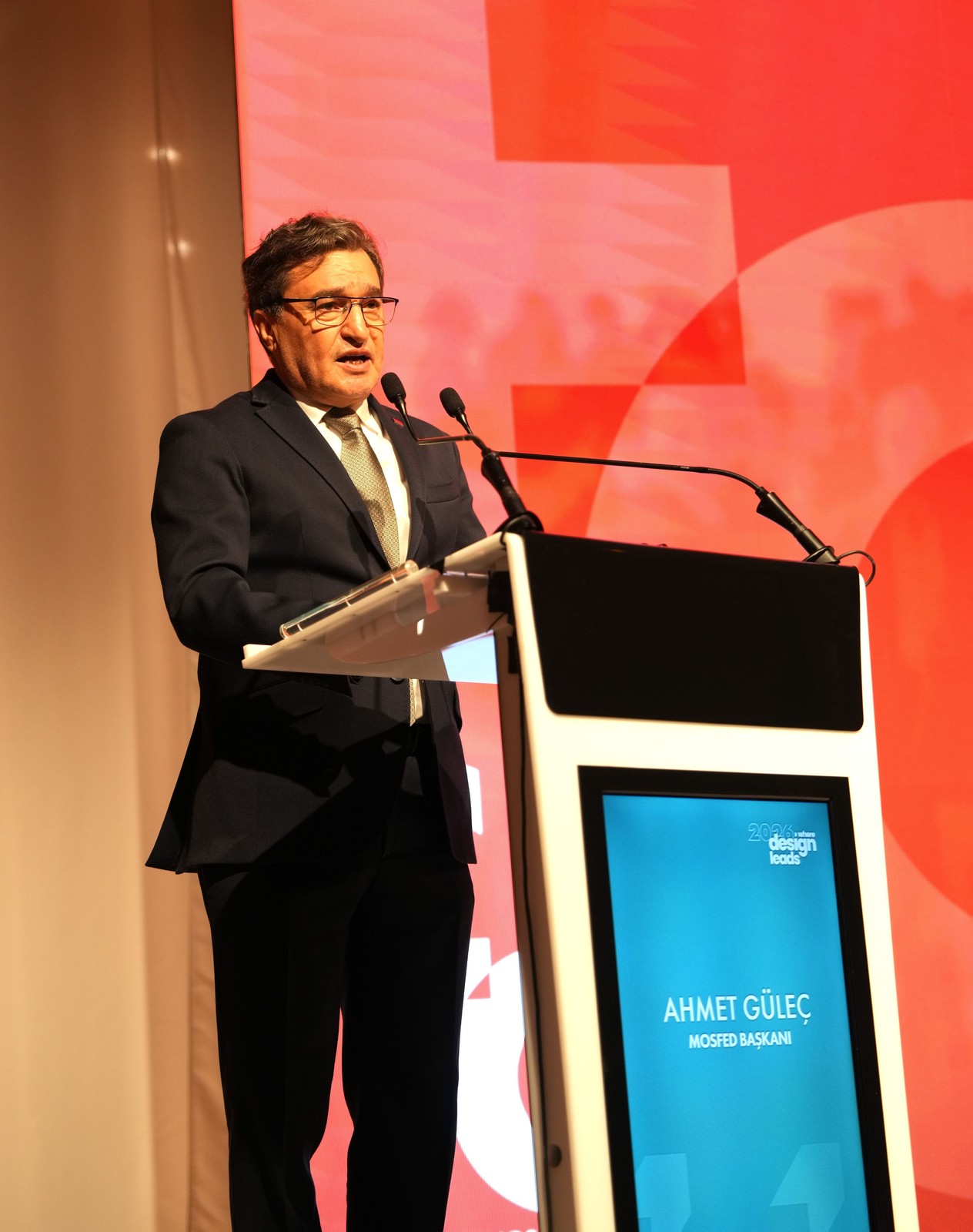 Kırmızı fonlu sahnede kürsüde konuşan MOSFED Başkanı Ahmet Güleç; kürsü ekranında adı ve unvanı yazıyor