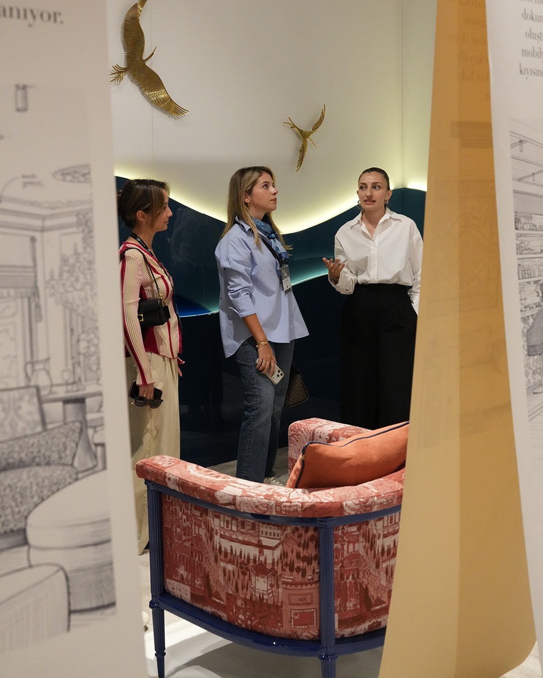 Fuar standında desenli kumaşla kaplı koltuğun ardında sohbet eden üç kadın, duvarda altın kuş figürleri